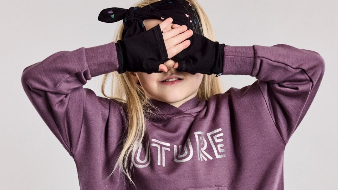 A child wearing a purple merino wool hoodie with 'FUTURE' printed on the front, black fingerless gloves, and a black headband with small colorful heart details, covering one eye against a light gray background.